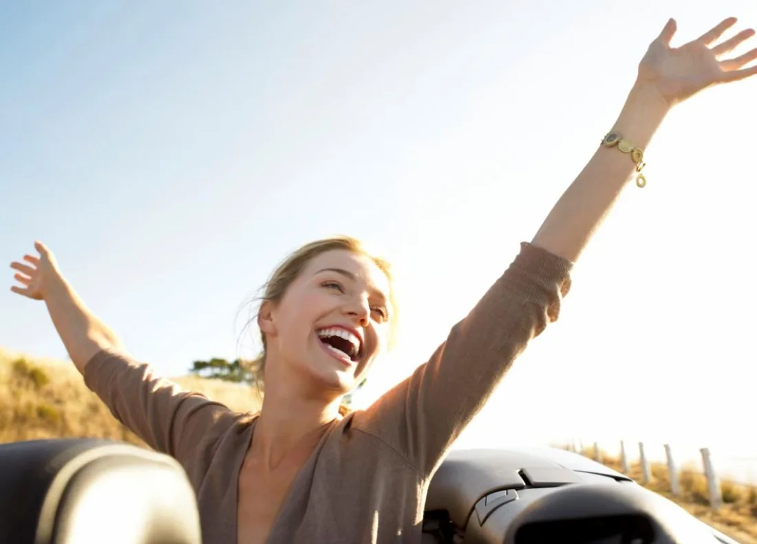 A woman laughing with both arms flung wide open in a convertible car under bright sunlight