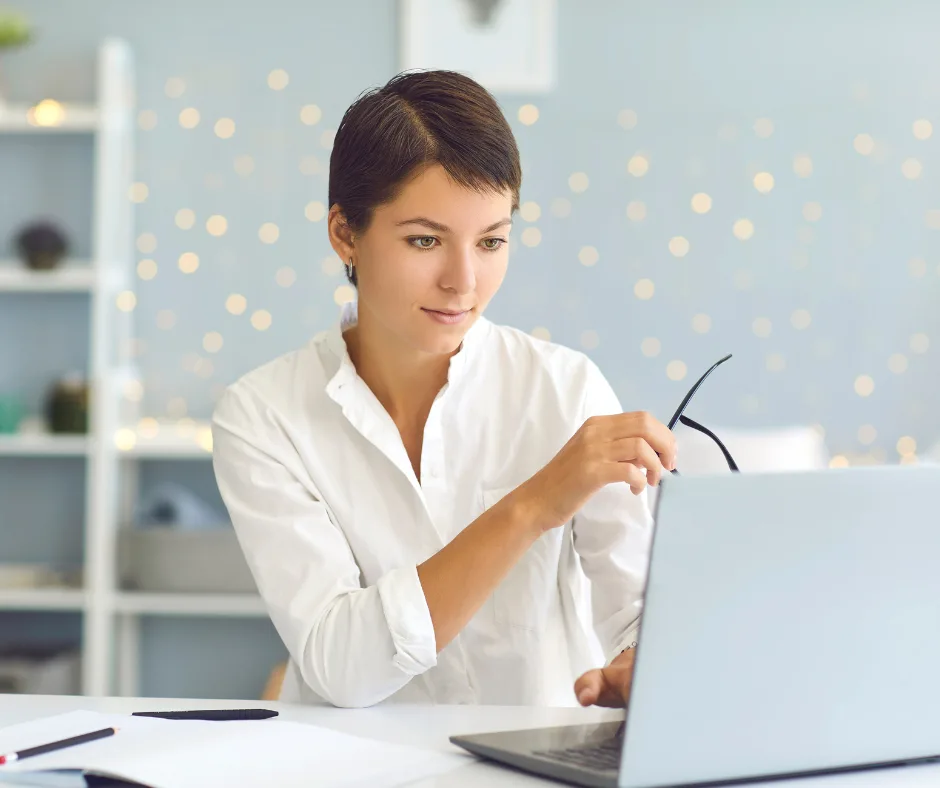 A focused professional pausing at her laptop, glasses in hand, in a bright home office