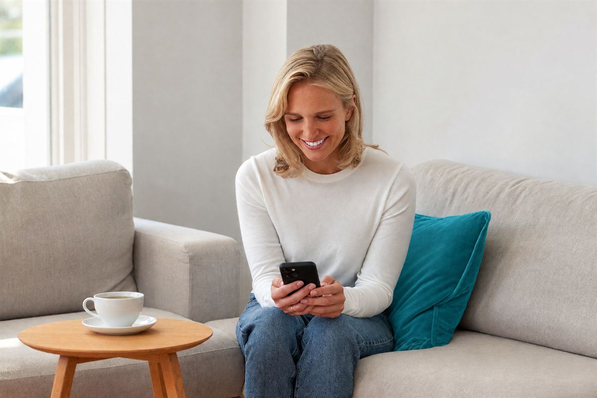 A woman on a light sofa beside a deep teal cushion, smiling as she answers a short quiz on her phone