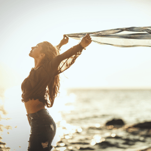 A woman on a beach at sunset holding a scarf up to the wind